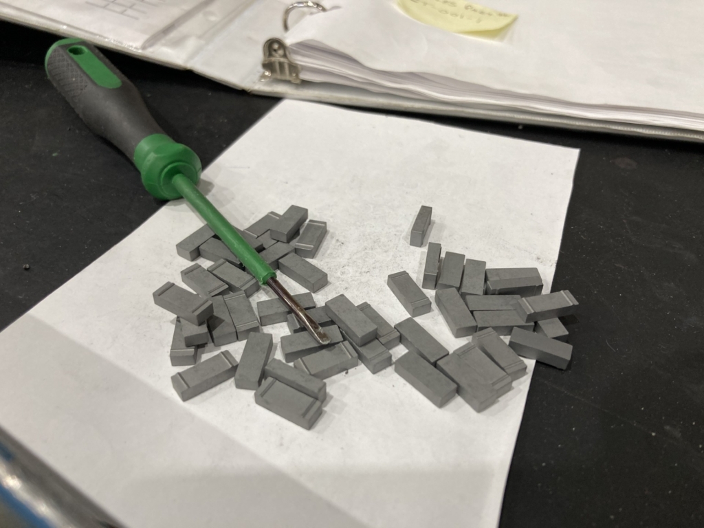 Rectangular tungsten carbide tiles sit atop a table waiting to be applied to a downhole tool component. A welding rod used to weld the tiles to the component sits beside.